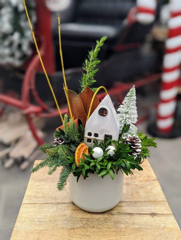 a small white ceramic filledw ith christmas greens, a faux white tree and white ceramic house accented with ornaments and orange slices.