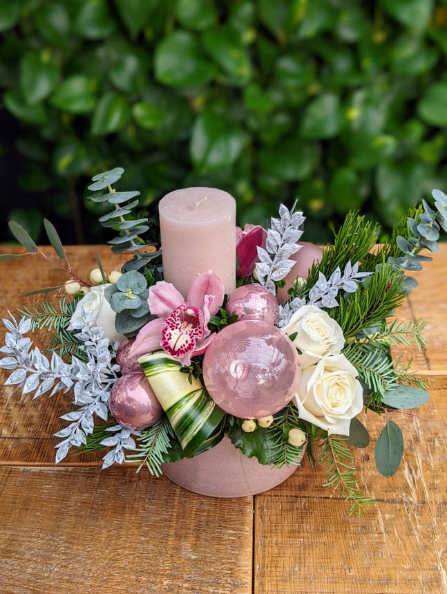 A pink glass container filled with white and pink florals and ornaments with a pink pillar candle