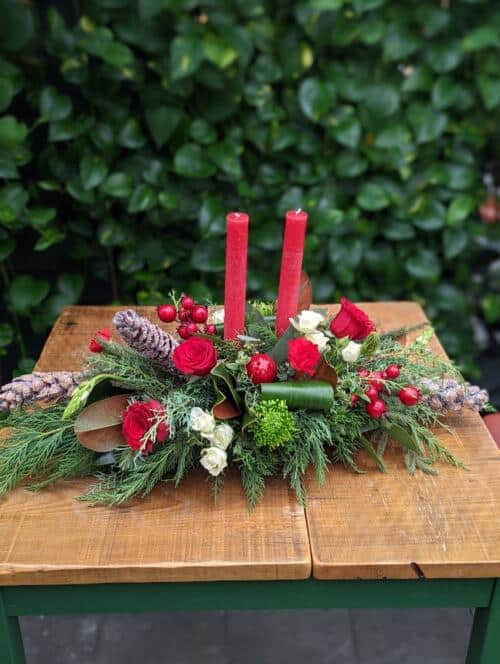 oblong centrepeice with 2 red tapered candles, red roses, white spray roses, red carnations, green mums, berries and pinecones