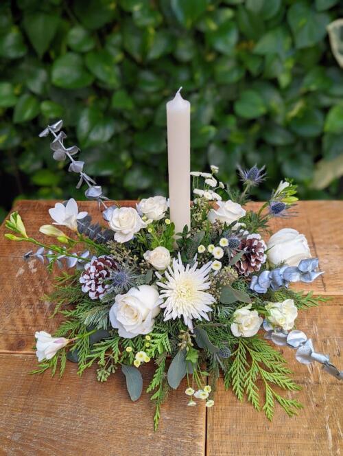 winter greenery with white flowers, silver greenery and a white candle. featruing white freesia, roses and mums, thistle and pinecones