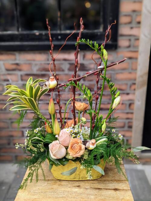 A yellow orval ceramic filled with peach, yellow and orange florals. featuring peach ranunculus, white freesia, orange tulipds, curly willow and more adorned with small birds.