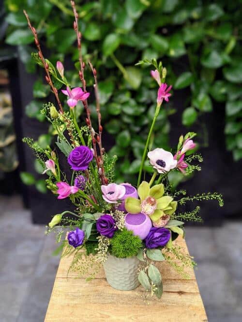 a light green textured ceramic filled with purple, green and white florals. an asymmetrical arrangement featuring anemones, purple freesia, purple lisianthus, green cymbidium orchids an d more