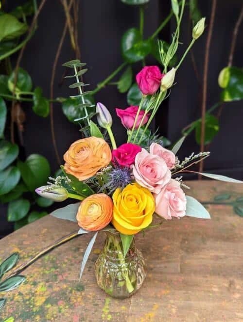 small glass vase featuring pink lisianthus, pink spray roses, peach ranunculus, purple tulips thistle and more.