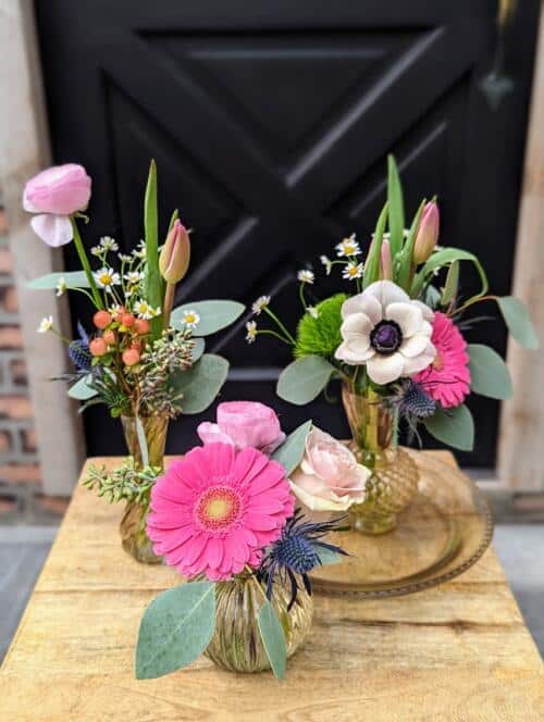 a trio of 3 glass vases on a glass plate overflowing with fresh spring blooms. featuring anemones, bright gerberas, tulips and more