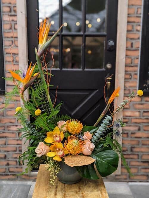 a large grey ceramic filled with orange, yellow and peach blooms accented with large tropical leaves. featuring shimmer roses, birds of paradise, pink cushion and more.