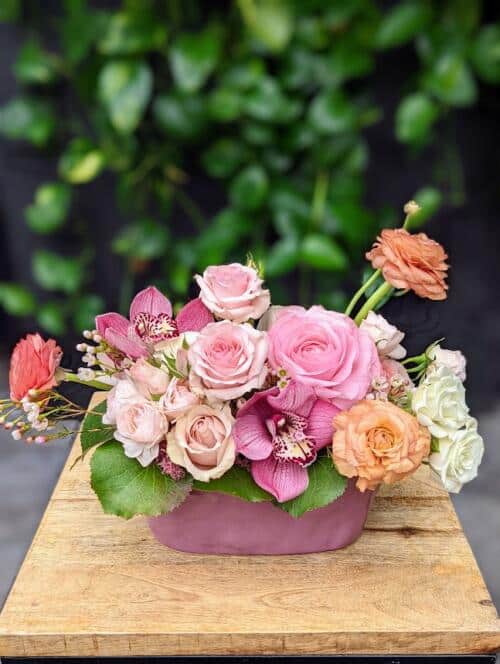 a purple oval ceramic filled with pink purple and peach blooms. featuring ranunculus, pink cymbidium orchids and roses.