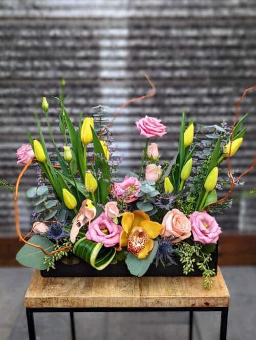 a large black metal container filled with spring flowers ready to burst. featuring yellow tulips, pink lisianthus, yellow cymbidium orchids, a variety of roses and greenery.