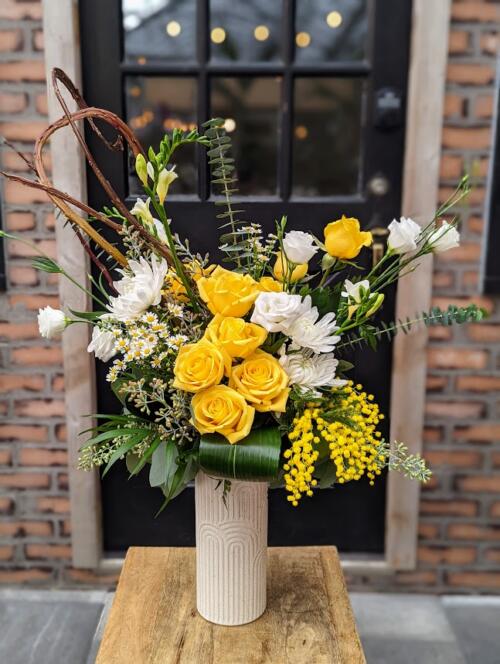 a white arch textured vase overflowing with yellow blooms. featuring yellow roses, chamomile, mimosa, white lisianthus and white mums. a bold pop of yellow.