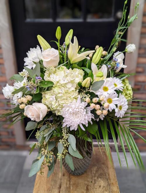 a clear textured vase filled with white and green flowers such as hydrangeas, lilies, mums and spray mums, accented with blue tweedia and seeded eucalyptus.