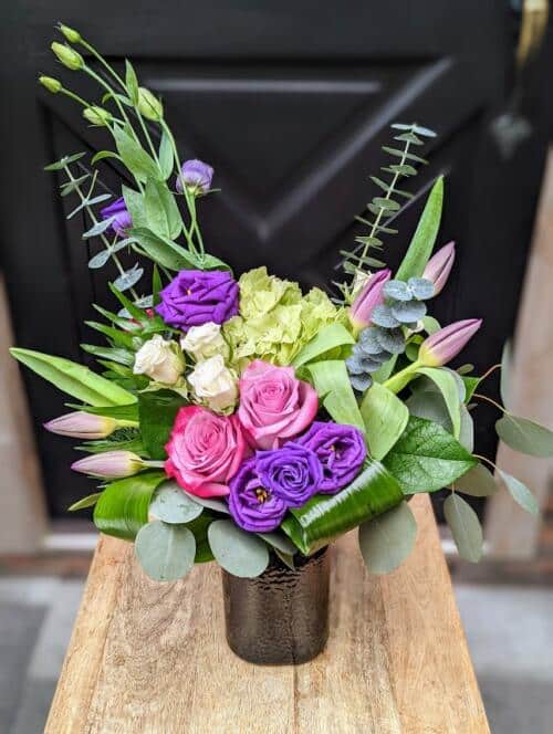 A coloured jar vase filled with purple and green blooms. featuring green hydrangeas, purple lisianthus and purple roses