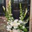 Tall white European floral arrangement with orchids, calla lilies, and garden roses in a cream vase in front of a black door