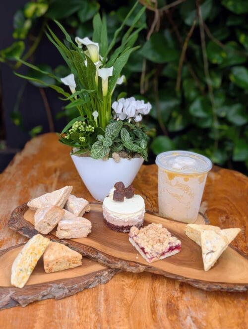 A rustic wooden board of freshly baked scones, a mini cheesecake, and a crumble bar beside a white pot planter of white calla lilies and cyclamen and an iced latte.