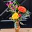 A petite amber glass bud vase holding a yellow rose, orange gerbera daisy, pink freesia, and blue thistle with greenery.