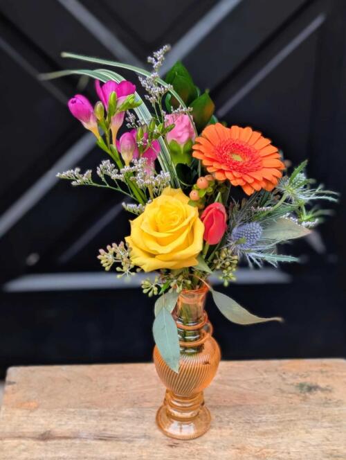 A petite amber glass bud vase holding a yellow rose, orange gerbera daisy, pink freesia, and blue thistle with greenery.