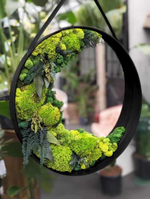 A round black-framed mirror edged with preserved moss, green chrysanthemums, and ferns.