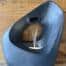 A dark slate-blue sculptural pour fountain with a smooth organic form and central circular opening, water cascading gently from a top spout through the hollow interior.