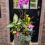 A wide view of a tall charcoal ribbed cylindrical pot on a wooden stand in front of a black door, with a magenta phalaenopsis orchid at the top, yellow calla lily, cream-speckled tropical foliage, orange kalanchoe, yellow mini roses, and trailing plants cascading down the front.