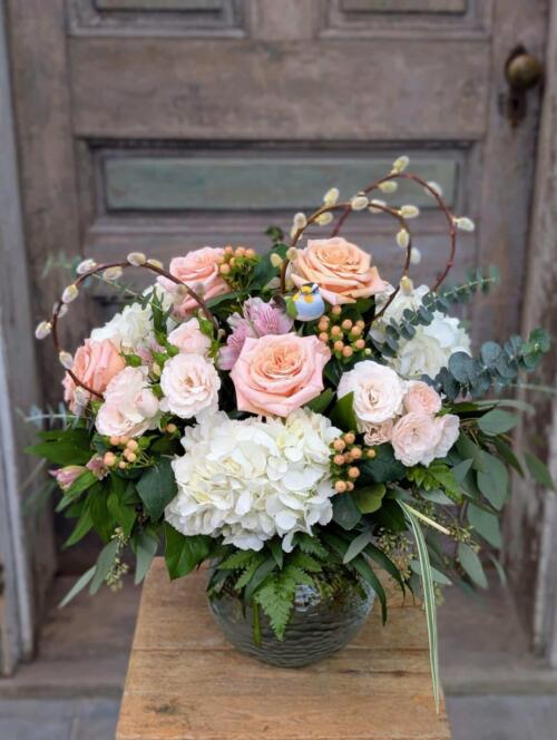 Mother's Nest floral arrangement in a glass bowl vase displayed in front of a rustic wooden door, featuring hydrangea, roses, and a bluebird figurine