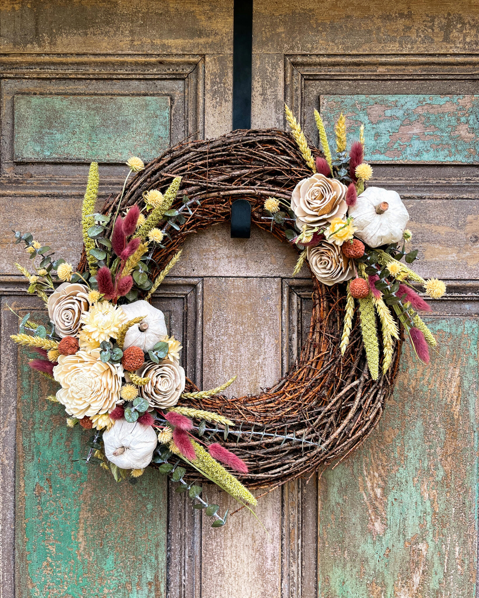 Autumn grapevine wreath decorated with sola wood roses, white mini pumpkins, dried bunny tail grasses, wheat, and craspedia, hanging on a weathered teal wooden door at The Watering Can.