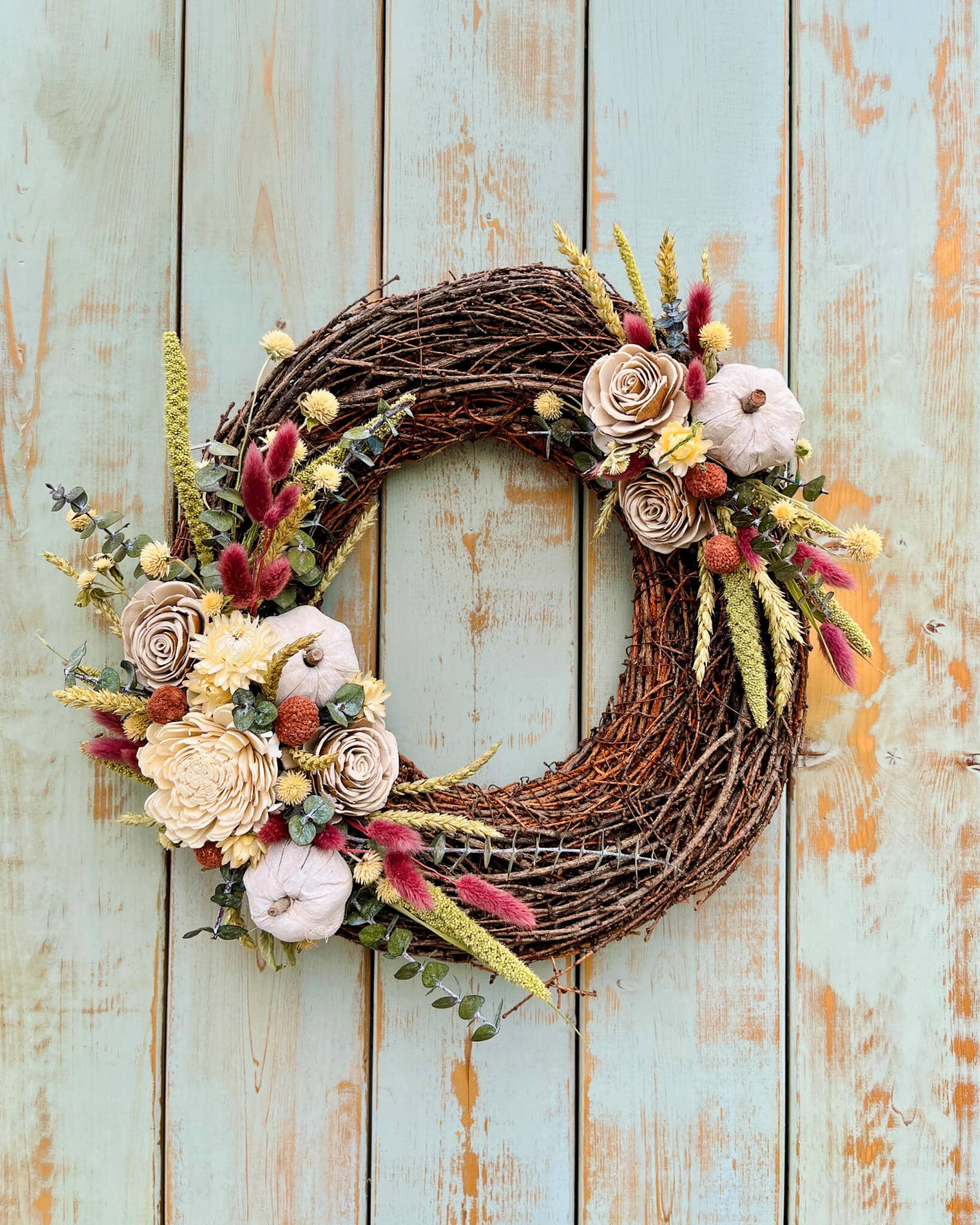 Autumn grapevine wreath with sola wood roses, cream mini pumpkins, dried grasses, wheat, and craspedia displayed against a weathered teal wood plank backdrop at The Watering Can.
