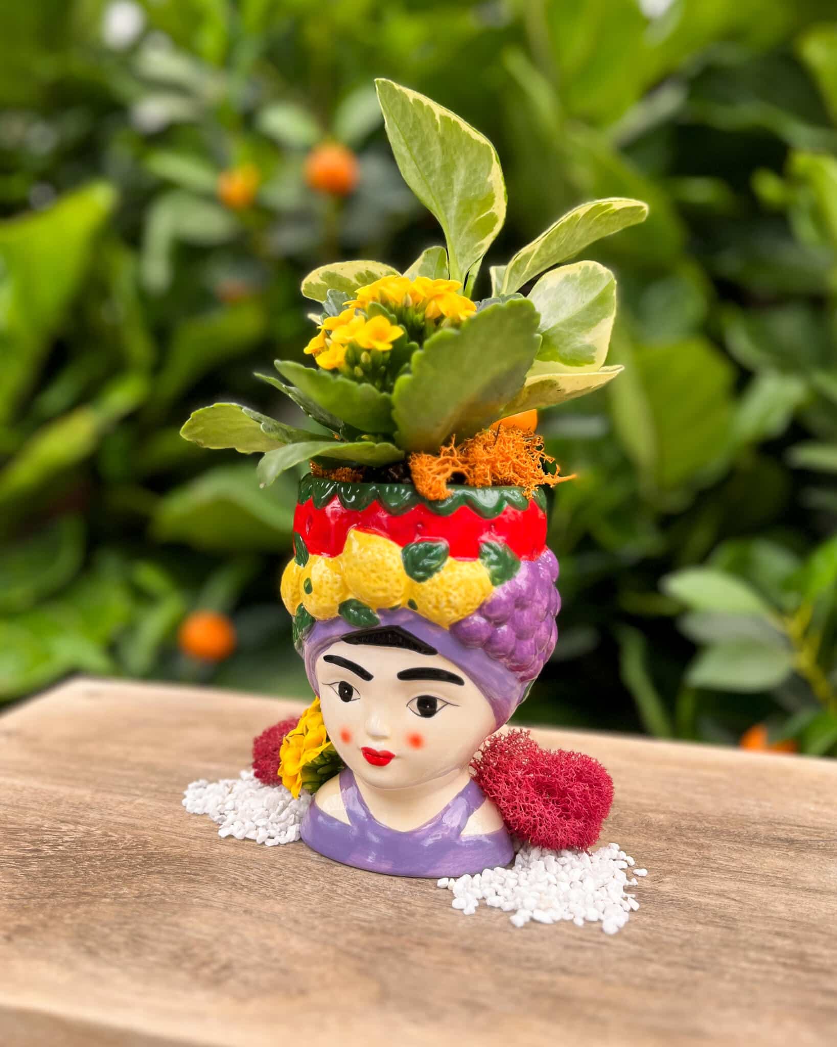 A whimsical ceramic lady-head planter with a lavender fruit-trimmed headwrap, planted with a bright yellow kalanchoe and variegated trailing foliage, displayed in the greenhouse.