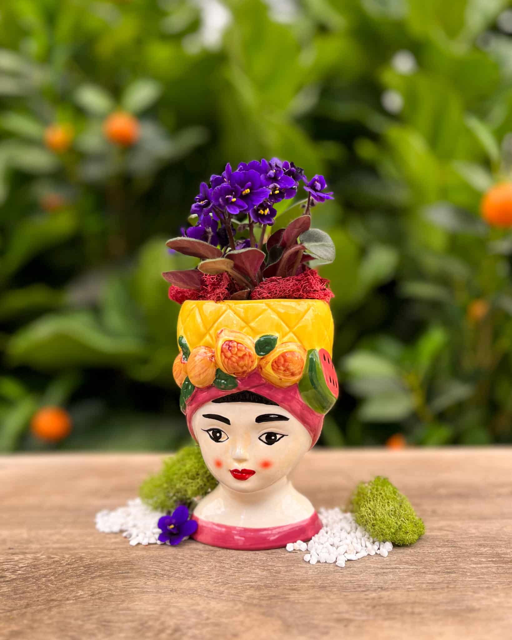 A whimsical ceramic lady-head planter wearing a fruit-trimmed yellow headwrap, planted with a purple African violet, displayed in the greenhouse.