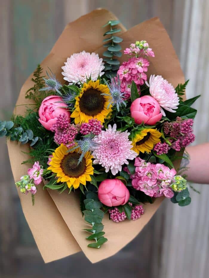 A hand-tied bouquet wrapped in kraft paper featuring bright sunflowers, pink peonies, pink chrysanthemums, stock and blue thistle accented with eucalyptus and fern.