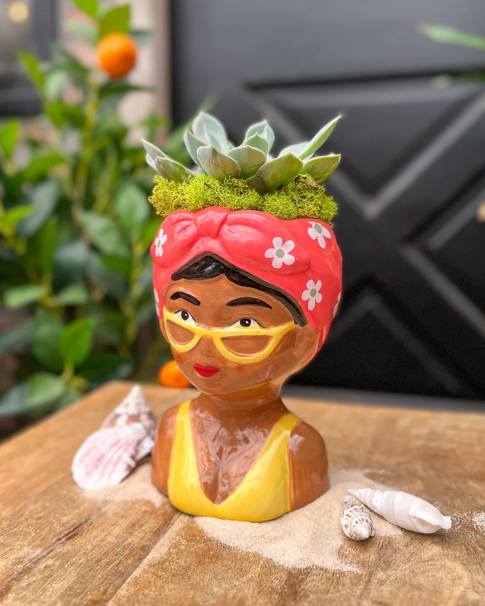 A ceramic lady-head bust planter with a pink floral headscarf, yellow glasses and a yellow dress, planted with echeveria succulents and moss.