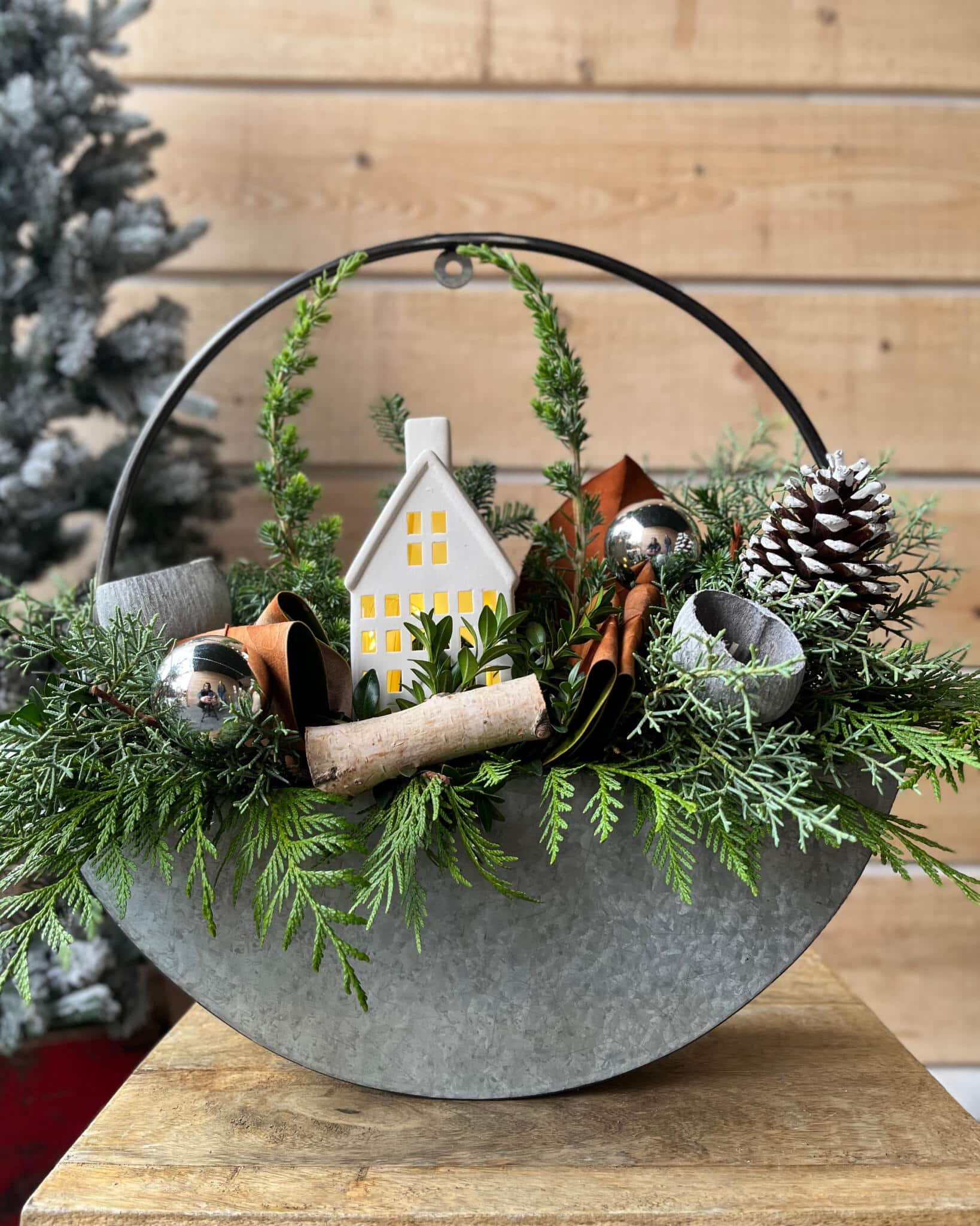 A round galvanized metal planter with a hoop handle filled with fresh cedar greenery, a small glowing white ceramic house, silver ornaments, a birch log and a pinecone, set on a wooden stand.