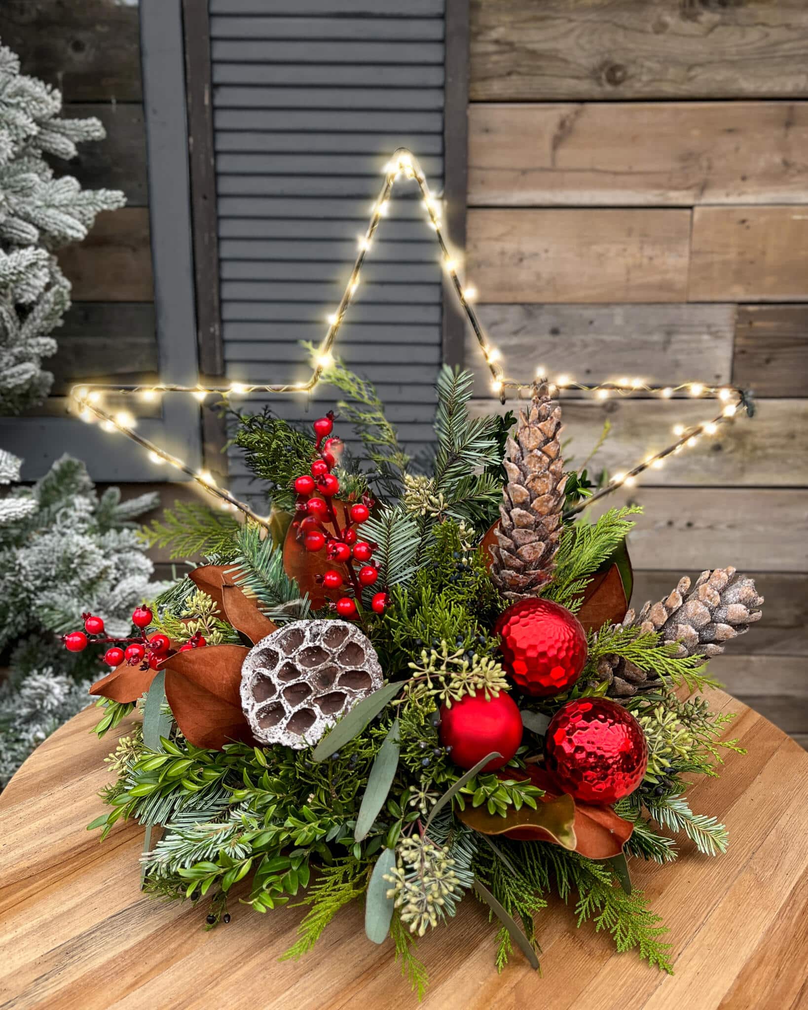 A festive winter centrepiece with a warm LED wire star rising from fresh cedar, pine and eucalyptus greenery, accented with red berries, pinecones, lotus pods and red ornaments, on a round wooden table.