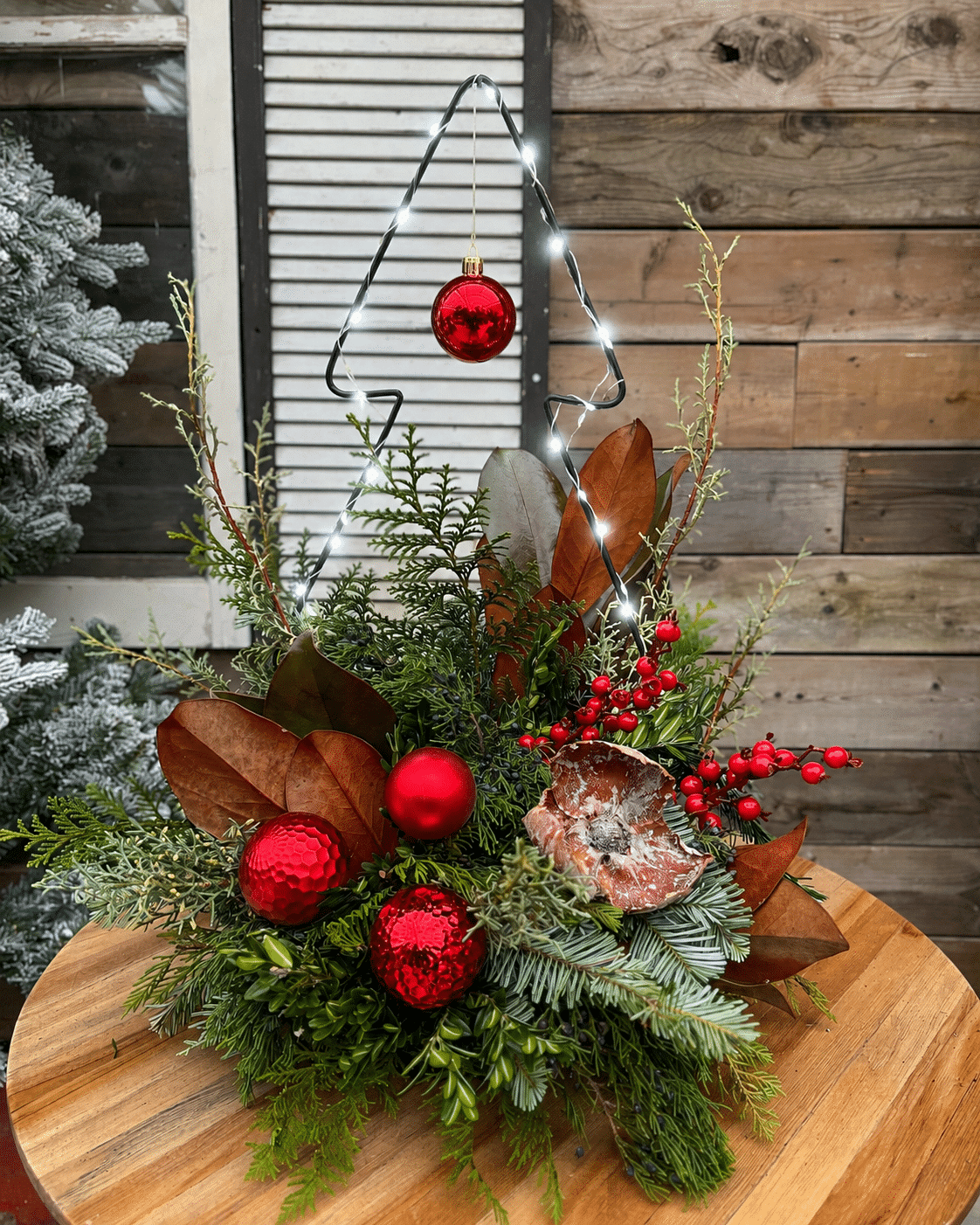 A festive winter porch planter filled with fresh evergreen boughs, red winter berries, pinecones, and red ornaments, glowing with warm string lights against a rustic wood backdrop.