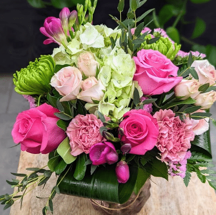 A hand-tied bouquet of hot pink and blush roses, white lisianthus, green hydrangea and pink carnations arranged in a leaf-lined glass vase.