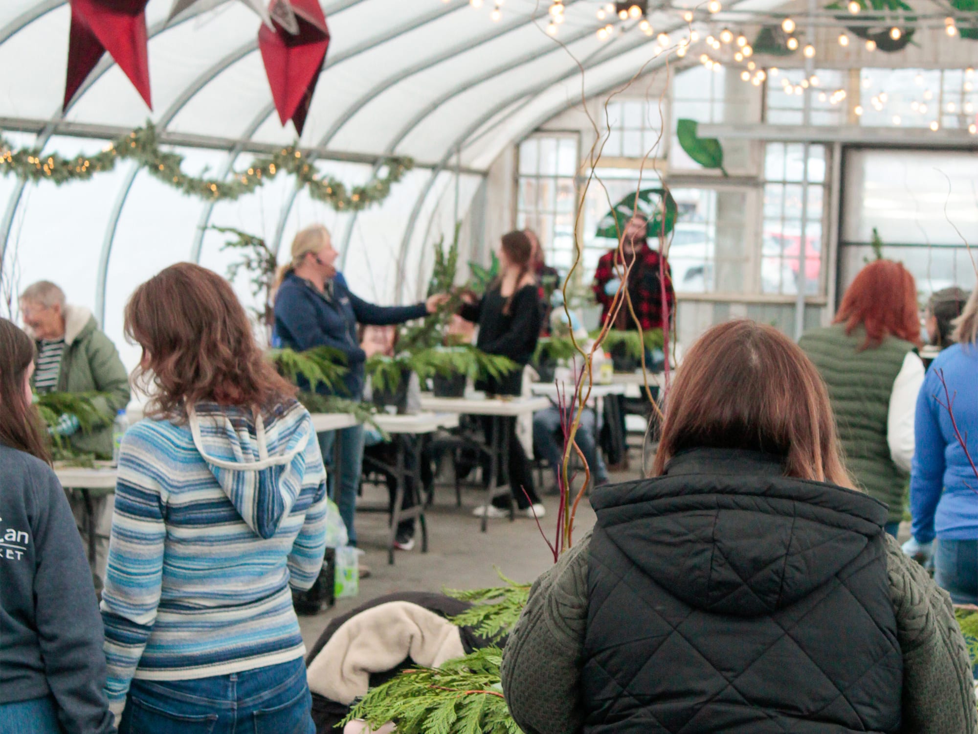 Guests gathered around tables inside a greenhouse workshop, arranging fresh evergreen boughs under twinkling string lights and red hanging decorations.